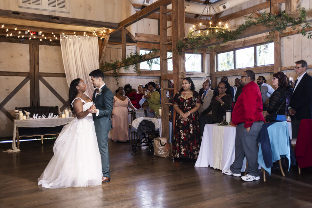 The couple shares their first dance in the barn’s open wooden interior, surrounded by smiling guests who watch from their tables; soft string lights overhead add a warm ambiance that defines The Farm Bakery and Events wedding as they sway together in the center of the room.