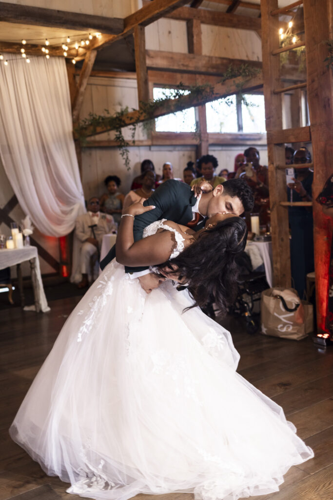 The groom dips the bride dramatically on the dance floor, kissing her as her gown fans out in a swirl of tulle. Guests gather around them, some cheering and others filming on their phones, with wooden beams and draped fabric framing the lively moment.