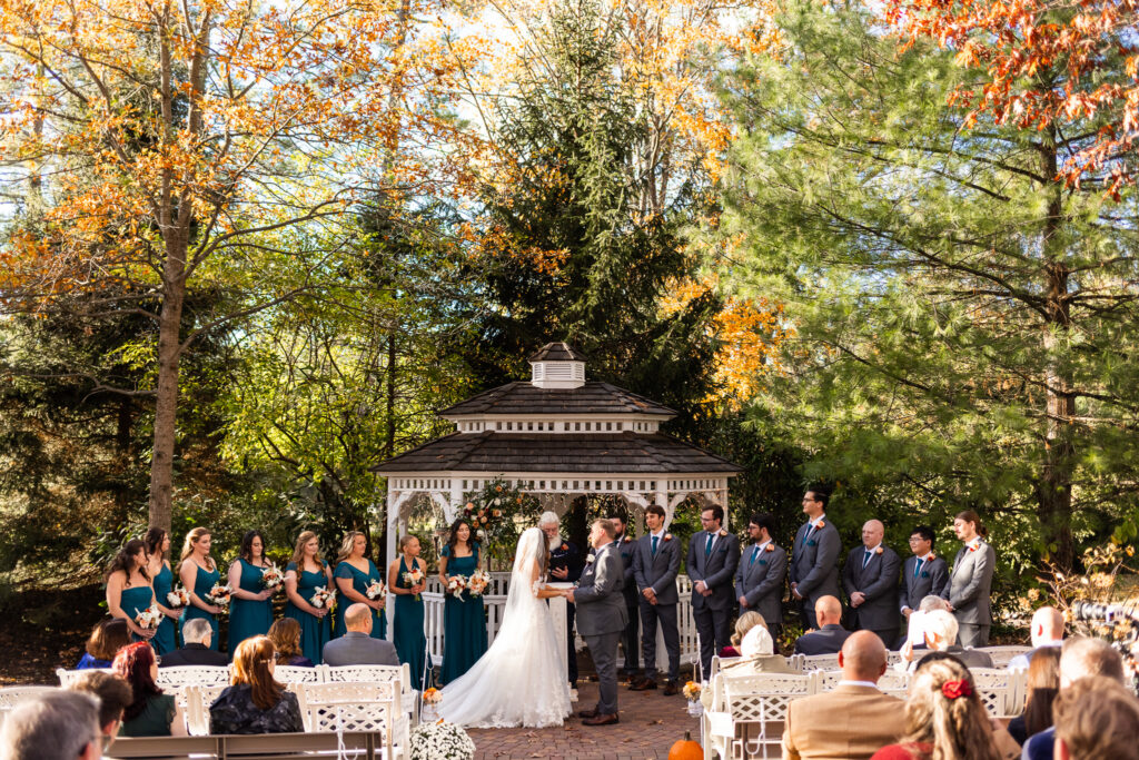 A couple stands at the center of an outdoor gazebo ceremony surrounded by tall fall trees with orange and green leaves, teal-clad bridesmaids to the left and groomsmen in gray suits to the right, while sunlight filters through the branches above; this scene, showing an aisle of seated guests watching the vows, clearly illustrates what time do weddings usually start during a crisp autumn afternoon.