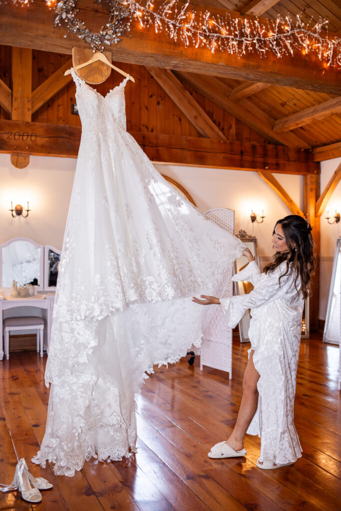 The bride stands barefoot in a cozy wooden bridal suite with vaulted beams overhead, gently lifting the lace hem of her wedding gown, which hangs from a wooden beam wrapped in twinkle lights. She looks up at the dress with a soft smile, her white robe trailing around her legs as warm light reflects off the polished floor. The ambiance of this moment captures the anticipation of a Pottstown Wedding in a charming rustic space where emotion and natural textures blend beautifully.