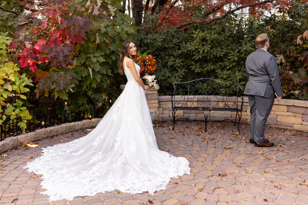 The bride stands behind the groom in a stone courtyard surrounded by lush fall foliage—deep reds, amber tones, and late-season greens. Her lace train fans out across the cobblestones as she smiles over her shoulder, bouquet in hand, just steps away from tapping him for their first look. In this peaceful outdoor scene, the golden hues of autumn amplify the emotional weight of their Pottstown Wedding, making the moment feel both intimate and cinematic.