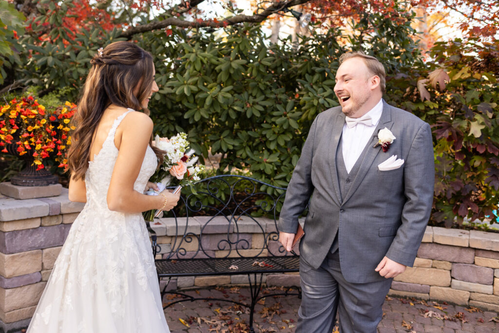 The groom turns toward the bride with a joyful, wide-open laugh as she steps closer holding her bouquet. They stand in front of a black wrought-iron bench surrounded by bushes brushed with red and orange leaves. Her expression is full of excitement, his full of disbelief and joy. The natural warmth of the setting highlights the heartfelt connection central to their Pottstown Wedding, making the scene glow with authentic emotion.