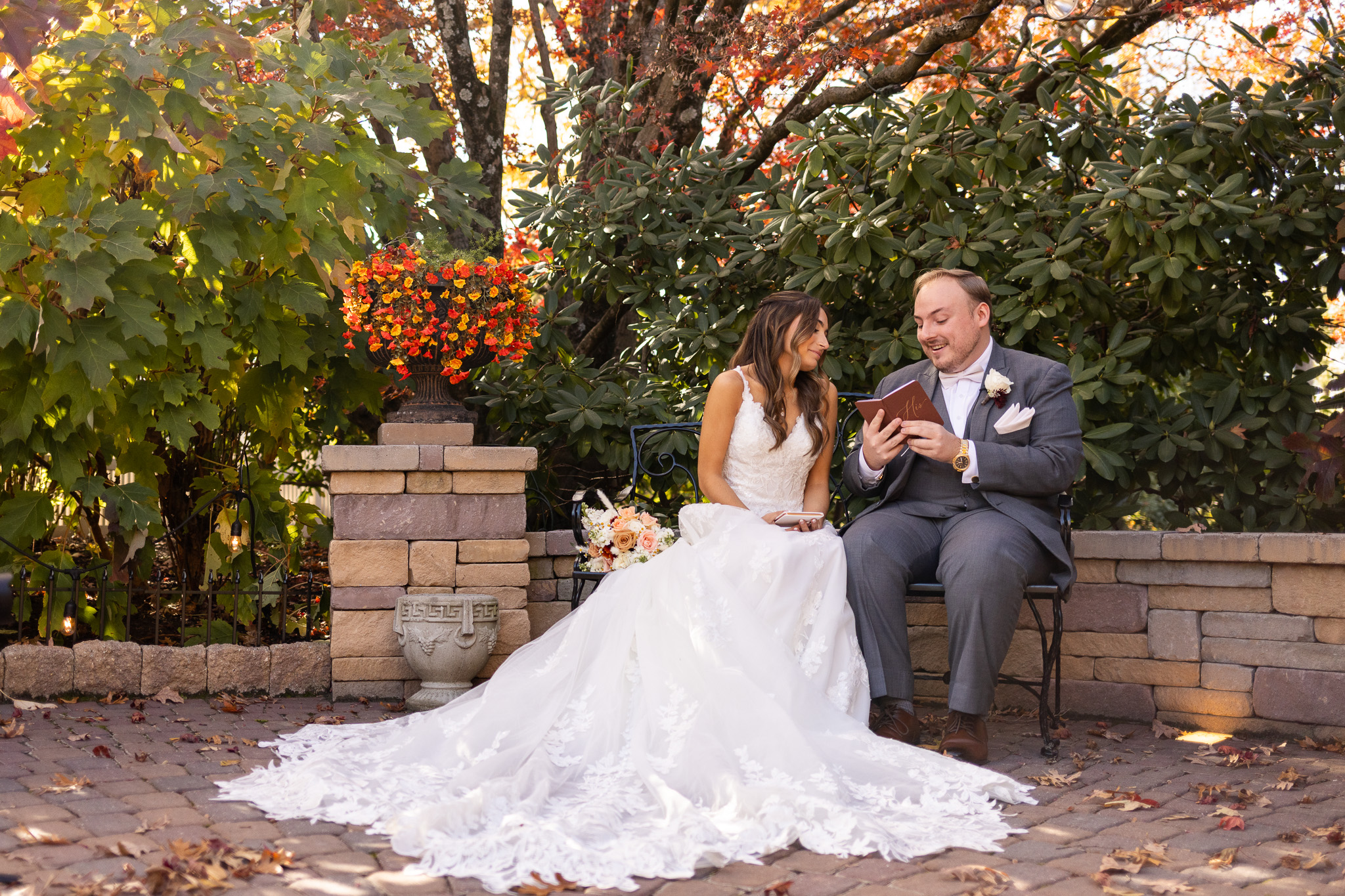 The bride and groom sit close together on a black wrought-iron bench in a quiet garden corner, surrounded by thick green shrubs and fiery autumn leaves. The groom reads from a leather notebook while the bride leans toward him with a soft smile, her lace train spilling across the stone patio. In the middle of the scene, the warm, intimate setting reflects the charm often found at wedding venues in Montgomery County, where natural landscaping creates pockets of calm for private moments. A bouquet of peach and cream flowers rests beside them on the bench.