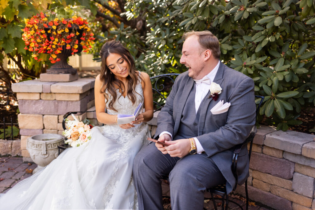 The bride and groom sit close together on a wrought-iron bench in a quiet garden nook, sunlight warming the stone wall behind them as she reads aloud from a small lavender vow book. Her lace gown spills across the brick pathway, and his gray suit catches the soft afternoon light as he leans in with a wide, joyful laugh. In the middle of this intimate moment, the peaceful landscaping—much like the settings you’d find at wedding venues in Montgomery County—creates a private pocket for their heartfelt vow exchange.