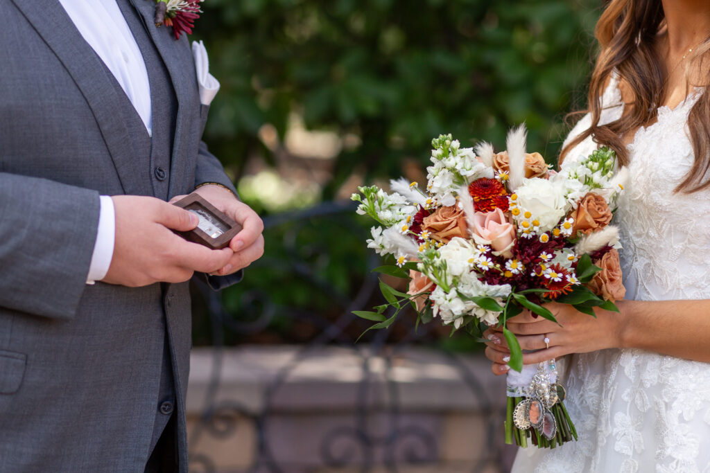 A tight crop shows the groom holding a wooden ring box with their wedding bands nestled inside while the bride stands beside him holding her bouquet of peach roses, white stock, burgundy mums, and tiny daisies. Her hand, with her engagement ring shining in the light, rests just above charms tied to the stems. In the middle of the frame, the elegant, natural backdrop echoes the refined outdoor spaces seen at wedding venues in Montgomery County, adding warmth to this detail moment.