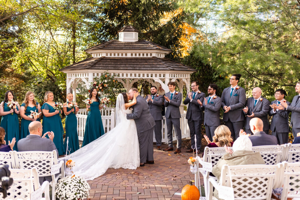 The bride and groom share their first kiss in front of the white gazebo, her long lace train fanned out behind them as the wedding party erupts in applause on both sides. Bridesmaids lift bouquets with excitement while groomsmen clap and cheer. Guests lean forward in their seats to witness the moment. At the center of this celebration, the beauty of the Meredith Manor venue elevates the kiss with its bright ceremony space and surrounding greenery.