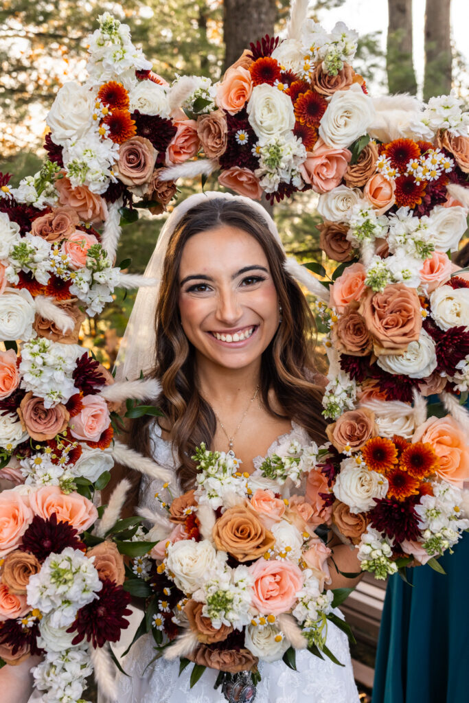 The bride poses with a radiant smile, framed tightly by an arch of florals held by her bridesmaids—soft peach roses, rust-colored mums, white stock, and fluffy pampas grass forming a vibrant halo around her face. Her veil drapes over her shoulders while she holds her bouquet close to her chest. This creative portrait style, often seen in the middle of galleries from wedding venues in Montgomery County, emphasizes color, texture, and joyful expression.