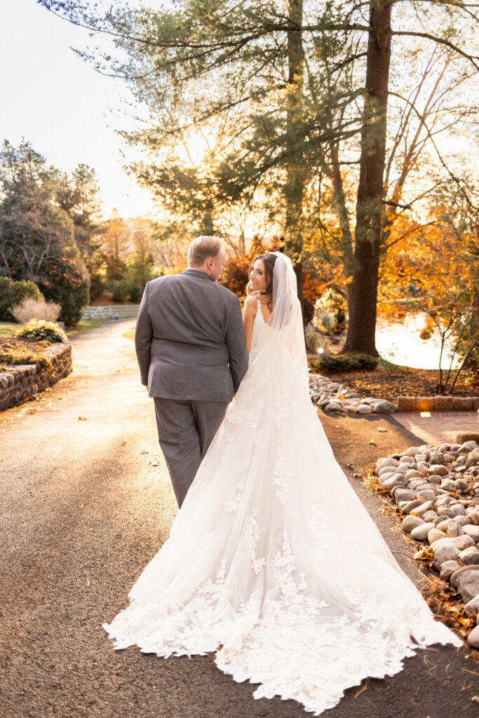 The couple walks down a gently curving pathway lined with stones and tall trees glowing orange in the setting sun. The bride’s long lace train trails behind her like a soft waterfall as she turns her head back toward the camera with a bright, joyful smile. The groom walks beside her, hands in his pockets, relaxed and content. The peaceful golden-hour landscape of the Pottstown wedding venue fills the middle of the scene with warm, romantic light.