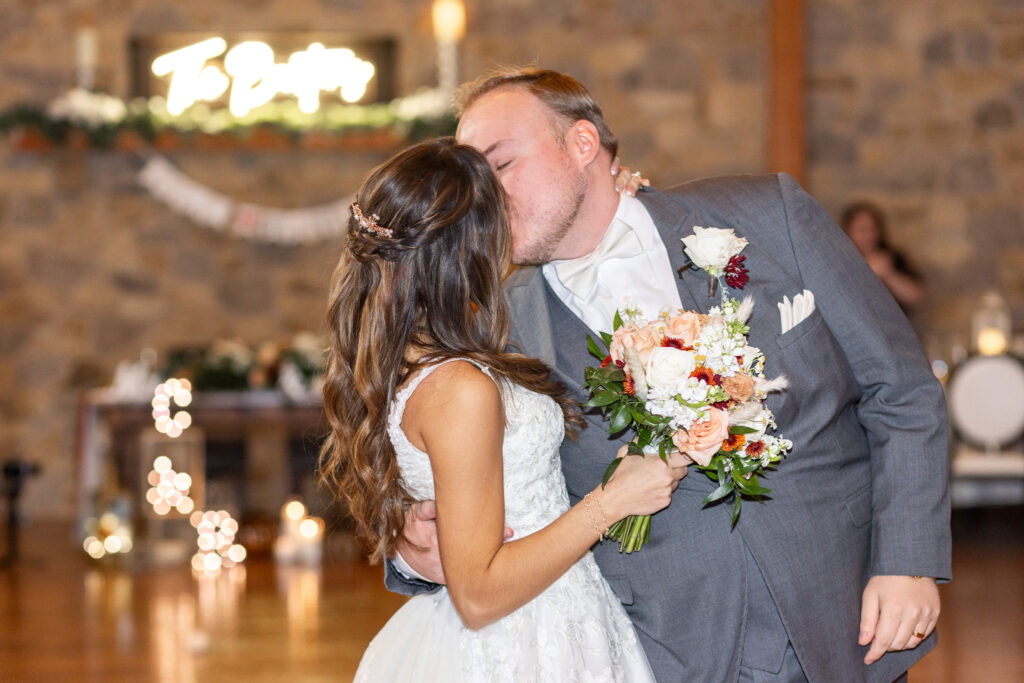 Inside a rustic stone-and-beam reception hall, the bride and groom share a kiss on the wooden dance floor, her bouquet of peach and ivory flowers held between them. Twinkling lights blur softly in the background near the sweetheart table. In the middle of the image, the cozy indoor ambiance evokes the welcoming style of wedding venues in Montgomery County used for fall celebrations.