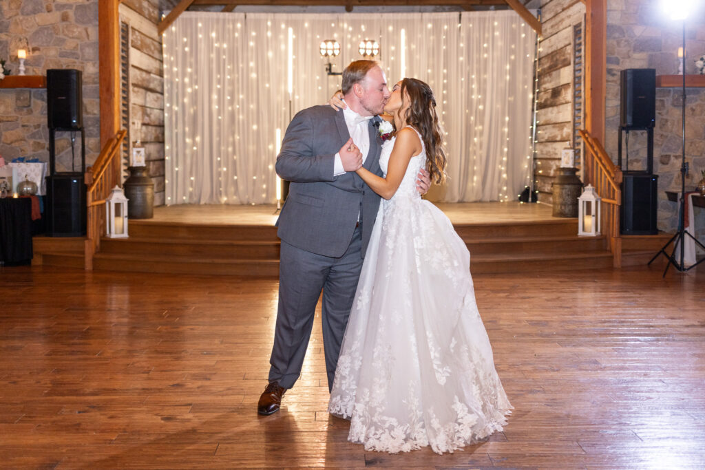 The bride and groom stand centered on a wide wooden dance floor, sharing a kiss beneath rows of string lights cascading down cream-colored drapery. Lanterns and wooden beams frame the space. In the middle area, the quiet, romantic setting feels similar to the intimate reception spaces offered by wedding venues in Montgomery County, ideal for private end-of-night portraits.