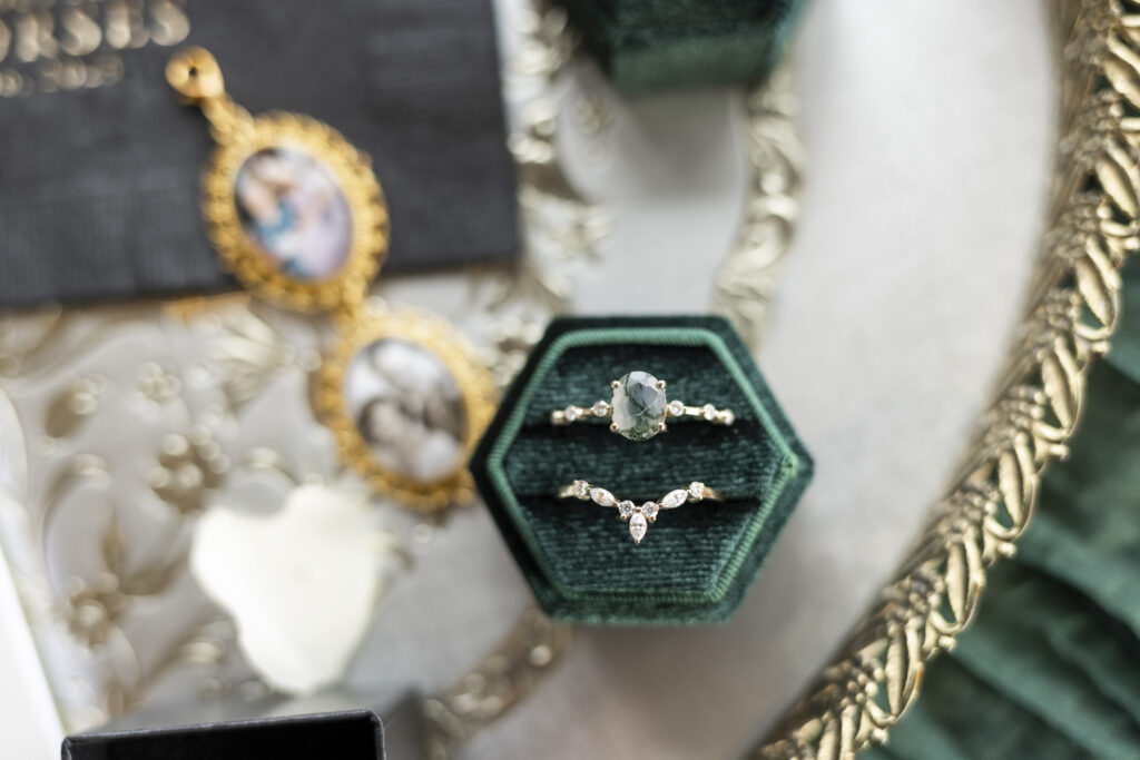 A close-up flat lay of two wedding rings resting inside a deep green velvet hexagon ring box, photographed on an ornate mirror tray, with gold-framed photo lockets and soft white details blurred in the background.