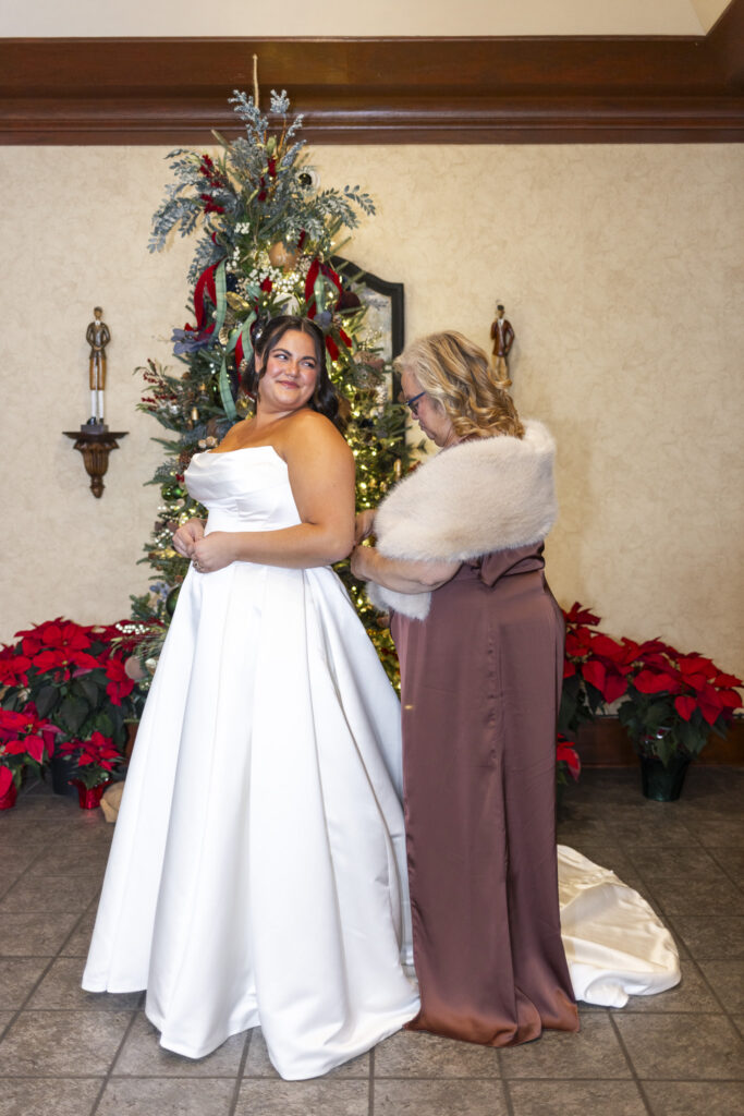 A bride in a strapless satin wedding gown stands in front of a decorated Christmas tree while an older woman gently buttons the back of her dress. The bride looks over her shoulder with a soft smile as poinsettias line the floor and warm indoor light fills the space at Lookout Mountain Golf Club, creating an intimate getting-ready moment.