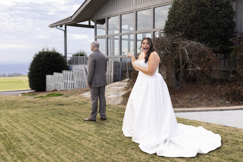 Outdoors on a grassy lawn, the bride stands behind her dad during a first look, pointing excitedly toward him with a wide, joyful expression. The groom faces away near the clubhouse building, and the open landscape of Lookout Mountain Golf Club stretches into the distance under an overcast sky.