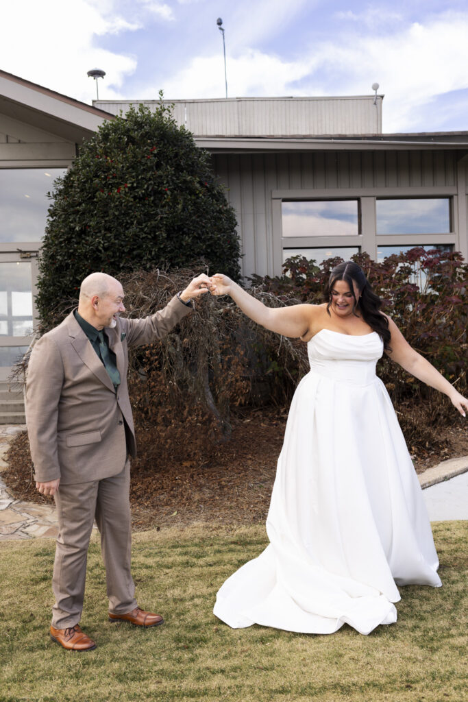 A bride in a strapless white wedding gown twirls joyfully while holding hands with an older man in a tan suit on the lawn of a lookout mountain wedding venue, her skirt lifting slightly as she smiles down at her movement with the building’s neutral exterior and winter landscaping behind them.