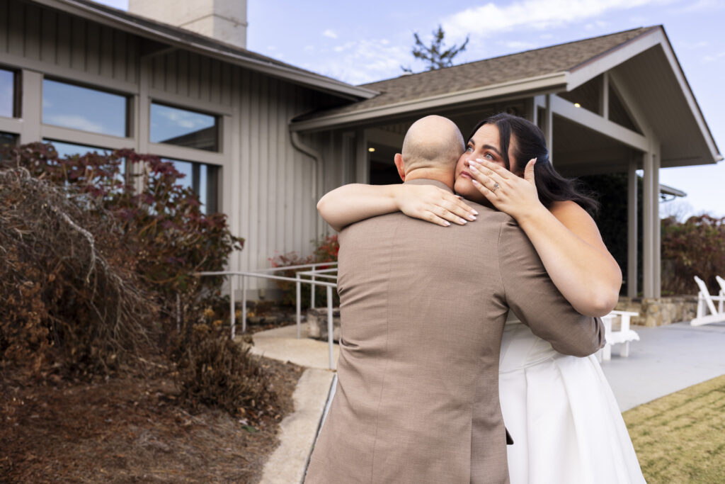 The bride embraces a bald man in a tan suit outside the clubhouse, her arms wrapped tightly around his shoulders as she wipes away tears with one hand. This was an emotional hug during the father-daughter first look