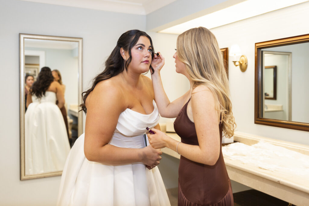 Indoors, a bridesmaid carefully applies mascara to the bride’s eyelashes as the bride leans forward slightly in her strapless satin gown. Mirrors reflect the scene behind them, and the softly lit powder room creates a calm, focused moment before the ceremony.