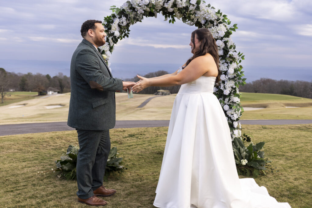 The couple stands facing each other beneath a circular floral arch, holding hands as they prepare to exchange vows. Rolling fairways and distant hills stretch behind them at the Lookout Mountain wedding venue, grounding the ceremony in open sky and landscape.