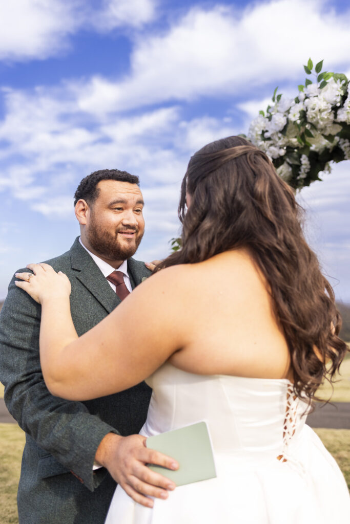 The couple faces each other during their ceremony at a Lookout Mountain wedding venue, the groom holding a small green vow book as he looks at the bride, with a floral arch and open sky behind them.