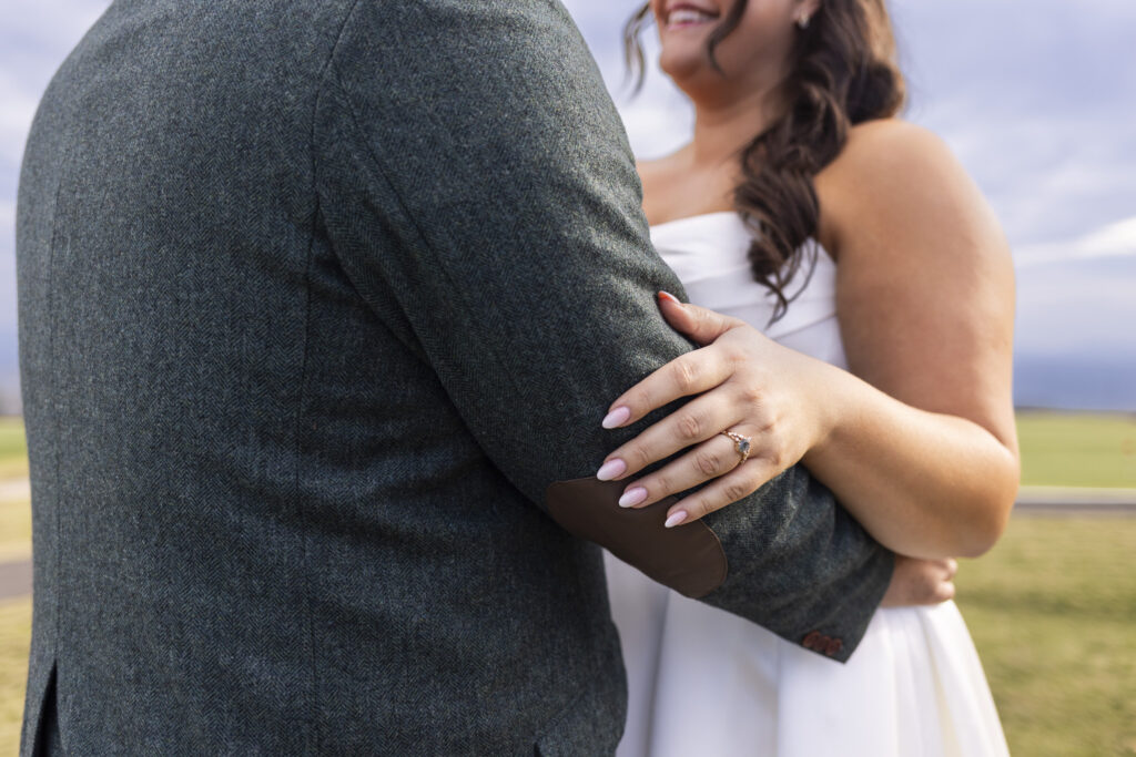 A close-up captures the bride’s left hand resting on the groom’s green suit jacket, highlighting her engagement ring and soft pink manicure.