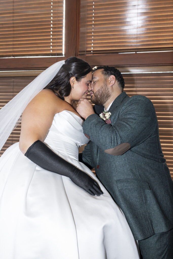 The bride sits atop a piano as the groom stand close, foreheads touching and eyes closed, as the groom gently cups the bride’s chin during an intimate portrait, warm light filtering through wooden blinds behind them.