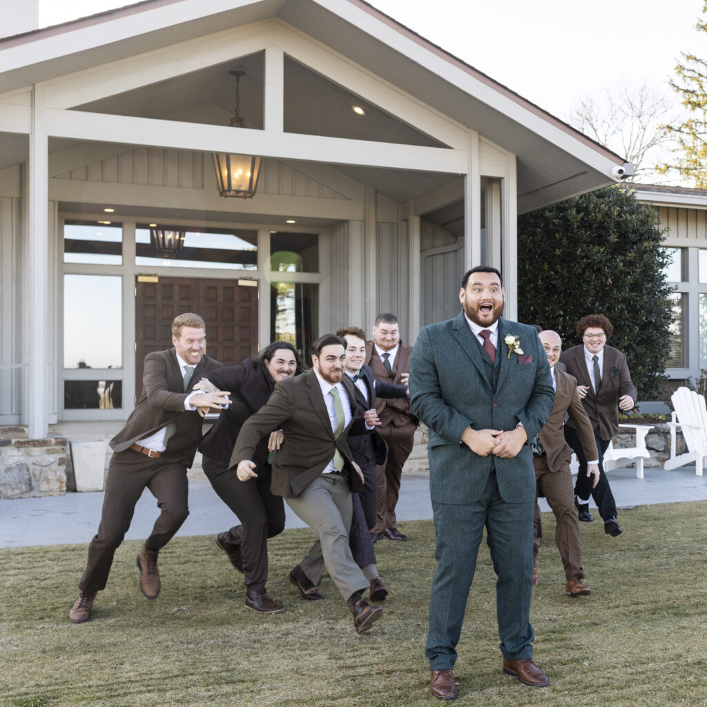 A group of groomsmen in coordinated suits runs playfully across the grass toward the groom, who stands in the foreground with a surprised expression. The modern clubhouse exterior of Lookout Mountain Golf Club sits behind them, emphasizing the lighthearted energy of this candid wedding party moment.