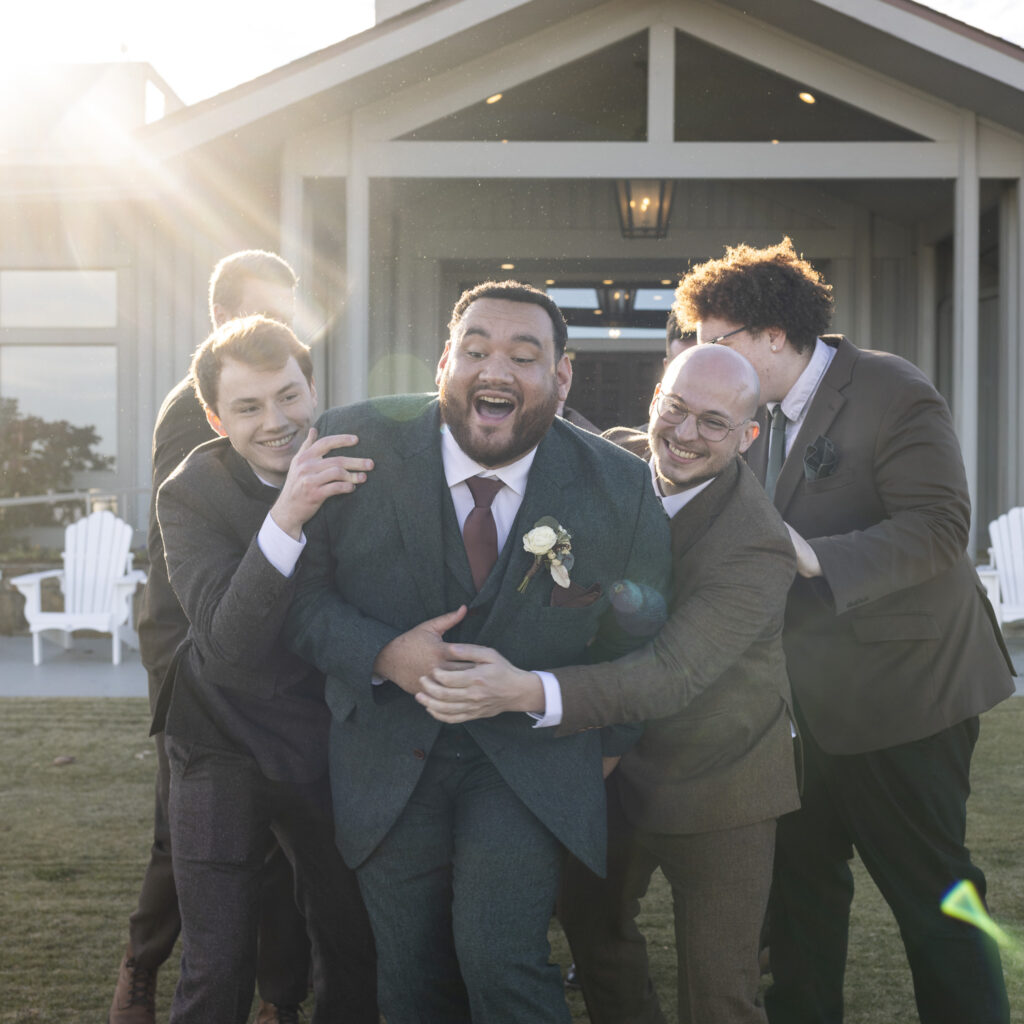 A joyful group portrait shows the groom laughing loudly as his groomsmen crowd around him outdoors, playfully grabbing his arms and shoulders while sunlight flares through the scene, all dressed in coordinated suits in front of the ceremony building just before the wedding.