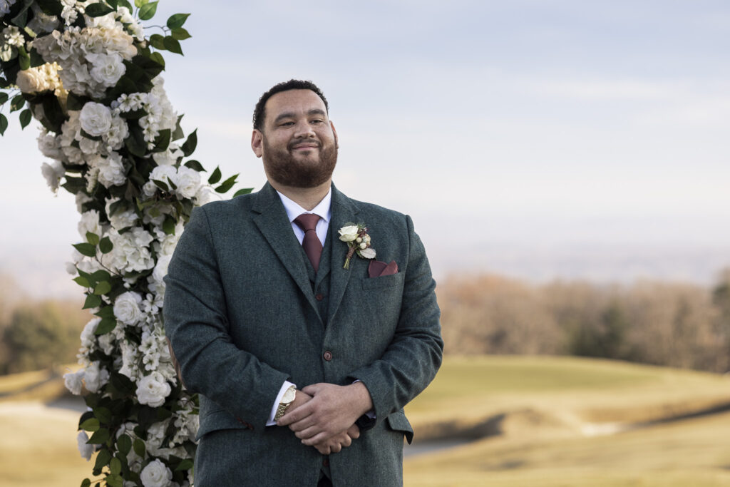 The groom stands alone beneath a white floral ceremony arch, hands clasped in front of him as rolling hills and muted winter trees stretch into the distance behind his dark green suit.