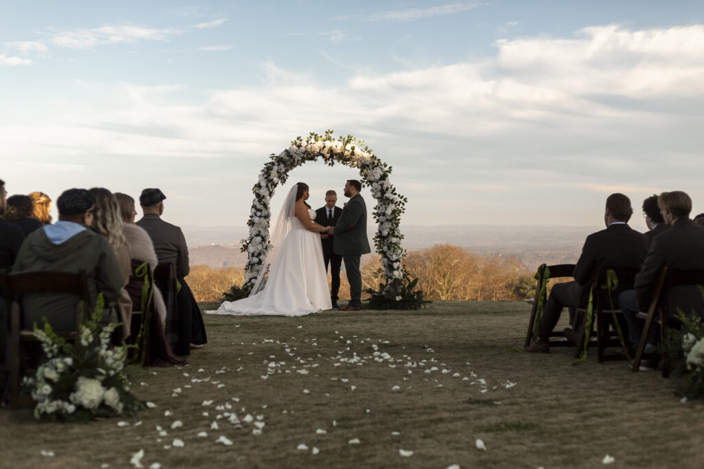 The couple stands facing each other during their outdoor ceremony beneath a large floral arch, holding hands as the officiant speaks. Guests sit on either side of the aisle, and flower petals scatter the grass, with sweeping mountain views visible beyond the ceremony space.