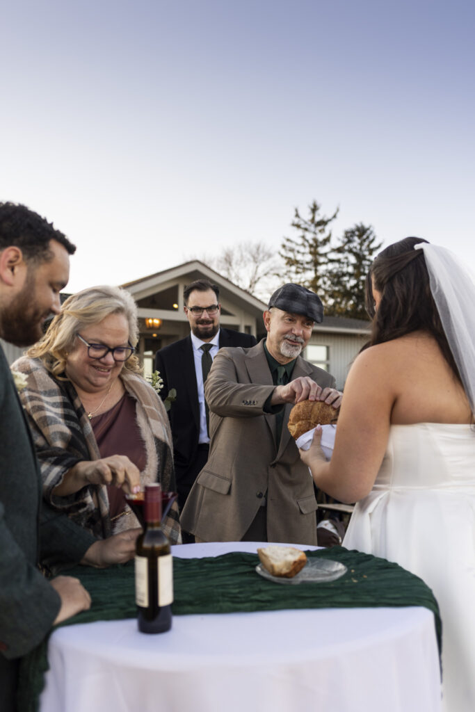 Communion takes place during an outdoor wedding ceremony, with family members gathered around a table as the bride holds a loaf of bread wrapped in a white cloth while a glass of wine is held by the groom for their guests to dip their bread into