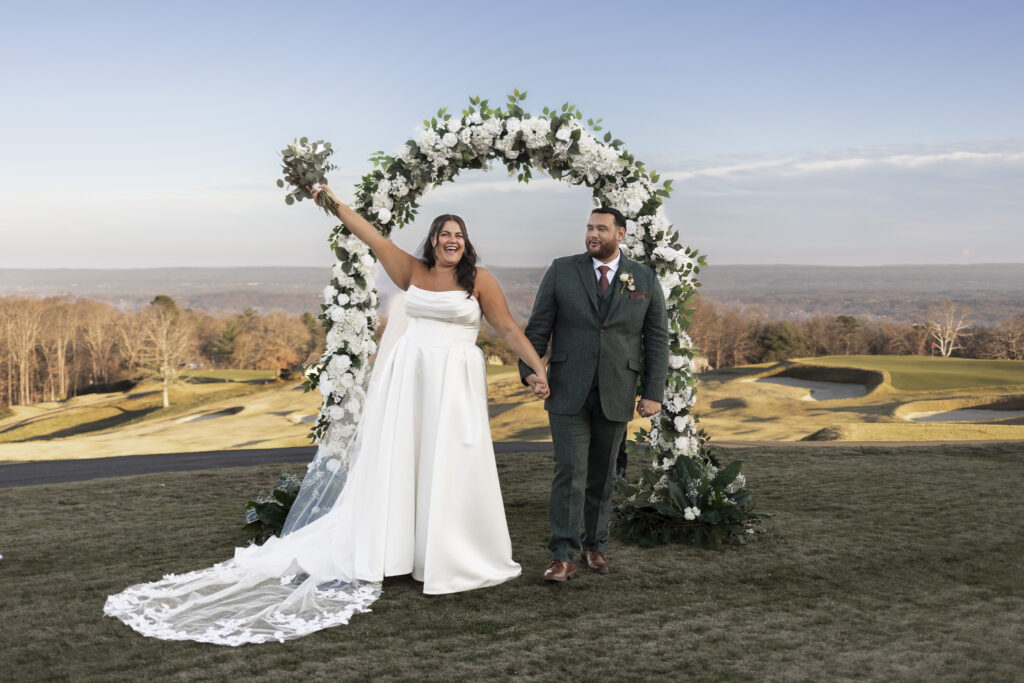 The newly married couple walks hand-in-hand beneath the ceremony arch at a lookout mountain wedding venue, the bride lifting her bouquet triumphantly as expansive mountain views frame the moment.