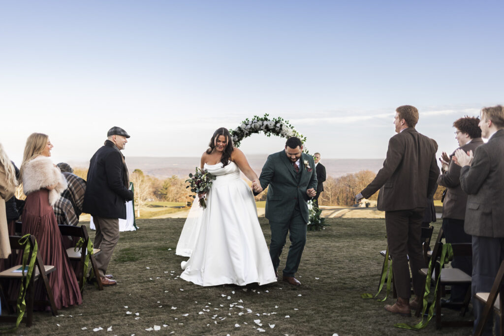 The newly married couple dances back up the aisle hand in hand, smiling as guests stand and applaud on either side. White flower petals scatter the grass, and the ceremony arch at the Lookout Mountain wedding venue rises behind them against a clear sky.