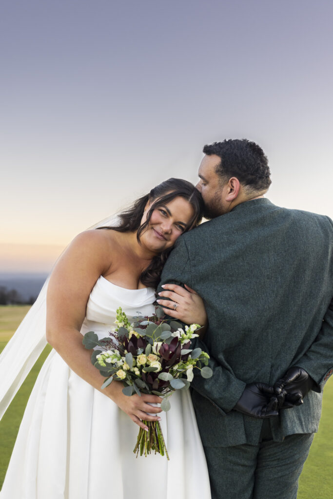The couple embraces during golden hour at a Lookout Mountain wedding venue, the bride resting her head on the groom’s shoulder while holding a bouquet of greenery and deep burgundy flowers against rolling hills in the distance.