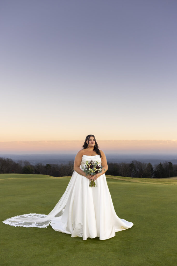 The bride stands alone on the green at dusk, holding her bouquet with her long veil trailing behind her across the grass. The horizon glows softly in the distance, and the open landscape creates a calm, expansive backdrop for this bridal portrait.