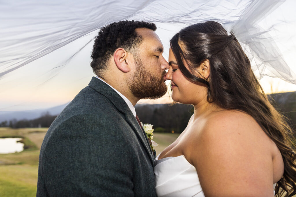 Under the bride’s flowing veil, the couple leans in with eyes closed, their noses touching as the fabric billows softly overhead. The rolling greens of the Lookout Mountain wedding venue fade into the background, keeping the focus on their quiet closeness.