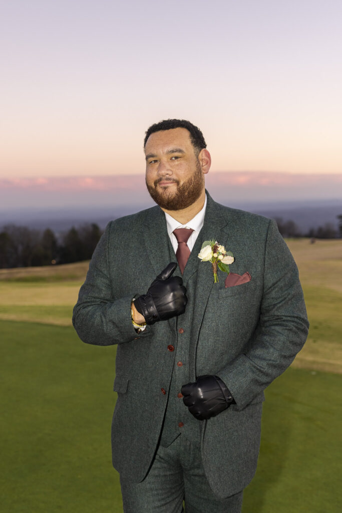 The groom stands alone on the golf course at sunset, wearing a green suit with leather gloves and a boutonniere pinned to his lapel. The pastel sky and expansive grounds create a striking, confident portrait.
