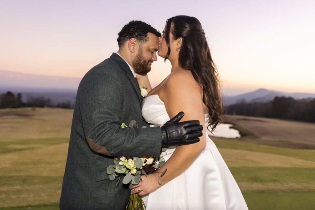 The bride kisses the groom’s forehead during sunset portraits at a Lookout Mountain wedding venue, her veil trailing behind her as the groom wraps his gloved arm around her waist with the mountains softly lit behind them.