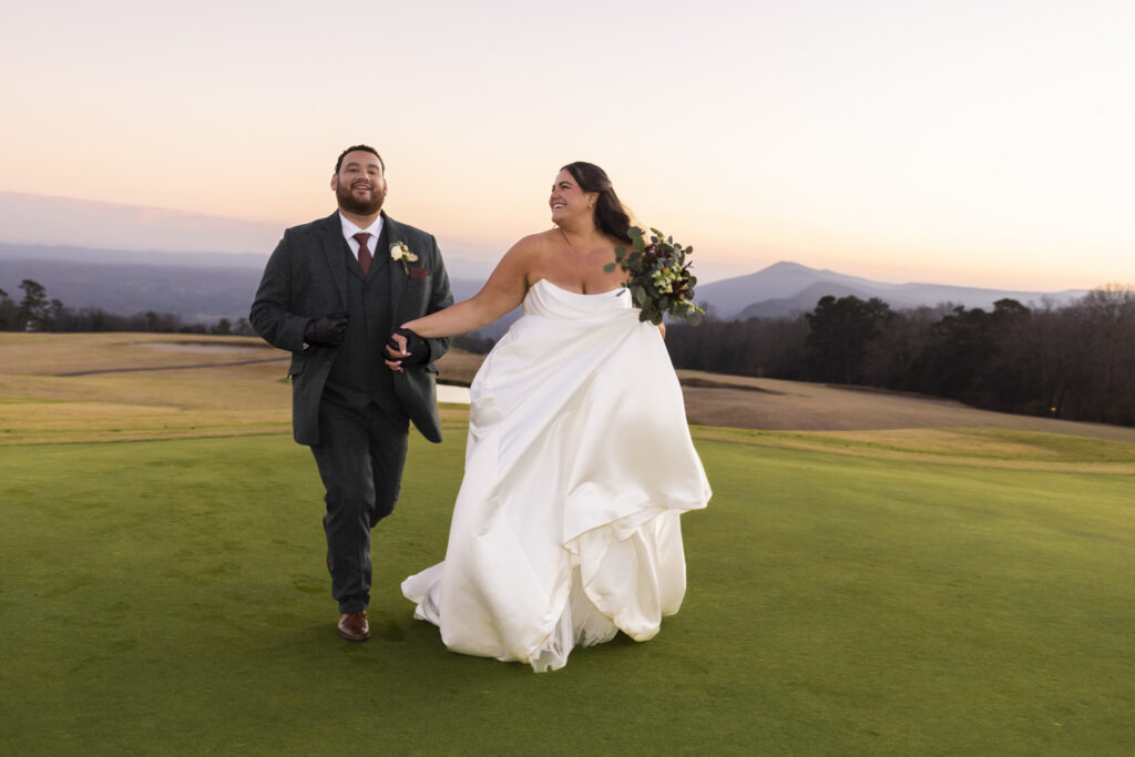 The newly married couple walks hand in hand across the golf course, laughing as the bride lifts her dress slightly and holds her bouquet. The groom smiles beside her, and the fading sunset over Lookout Mountain Golf Club adds warmth and motion to the scene.
