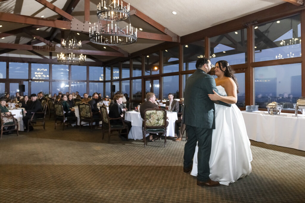 Inside the reception space, the couple shares their first dance surrounded by seated guests at round tables. Large windows reveal the darkening sky outside, while chandeliers glow overhead at Lookout Mountain Golf Club, capturing a cozy, celebratory end to the wedding day.