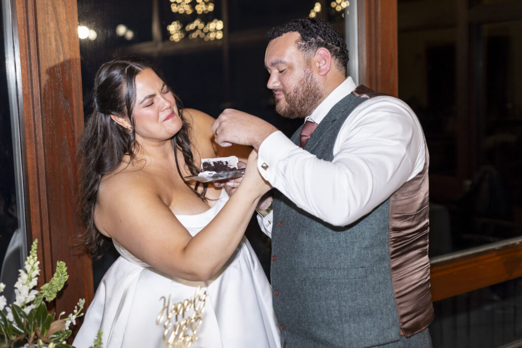 The bride feeds the groom a bite of chocolate cake during their cake cutting at a Lookout Mountain wedding venue, both smiling as they stand close together near a window with soft lights glowing outside.