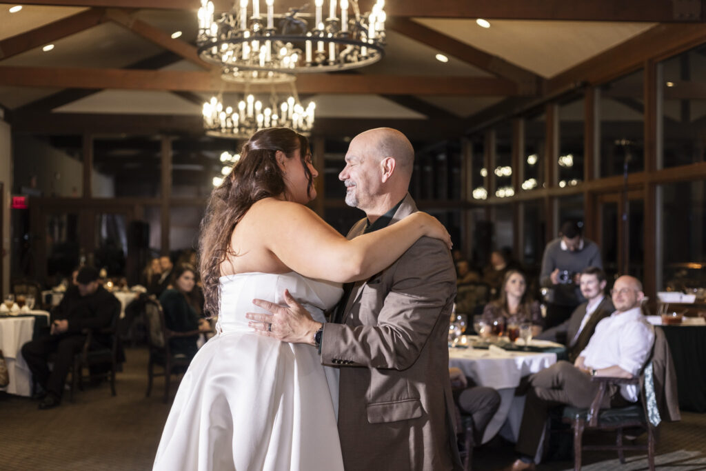 The bride dances closely with an older man during a parent dance, her arms around his shoulders as they share a quiet smile beneath chandelier light at the Lookout Mountain Golf Club.