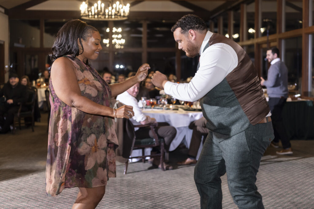 The groom dances with his mom on the reception dance floor at a Lookout Mountain wedding venue, both mid-step and smiling as seated guests watch from nearby tables under warm chandelier light.
