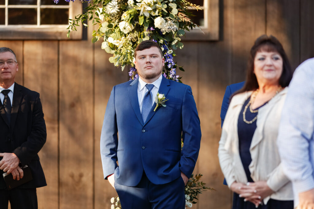 The groom stands beneath a floral ceremony arch wearing a blue wedding suit and boutonniere, hands in his pockets as he waits during the ceremony at coffey barn, with family members standing nearby and the wooden barn wall behind him.