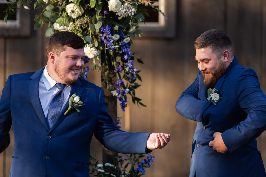 Two groomsmen in matching blue suits share a playful moment beneath the floral ceremony arch at the coffey barn, smiling as the best man searches for the rings in his jacket pocket