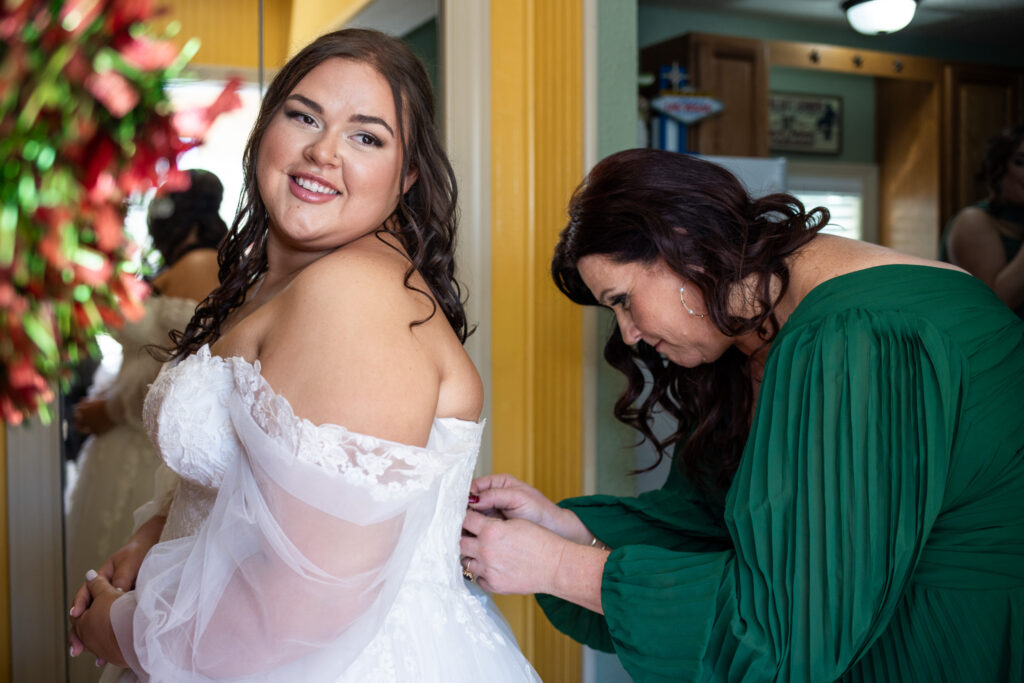 The bride smiles over her shoulder as a bridesmaid fastens the back of her lace wedding dress during getting ready. The bride wears an off-the-shoulder gown with sheer sleeves, while the bridesmaid in a green dress focuses on the buttons, capturing a tender, behind-the-scenes wedding moment.