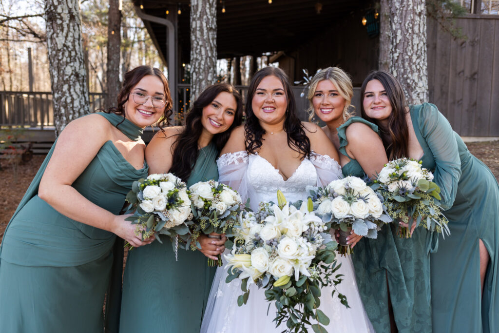 The bride stands at the center of her wedding party, surrounded by bridesmaids in coordinated green dresses holding white and greenery bouquets. Everyone leans in close, smiling toward the camera, capturing a joyful bridal party portrait before the ceremony.