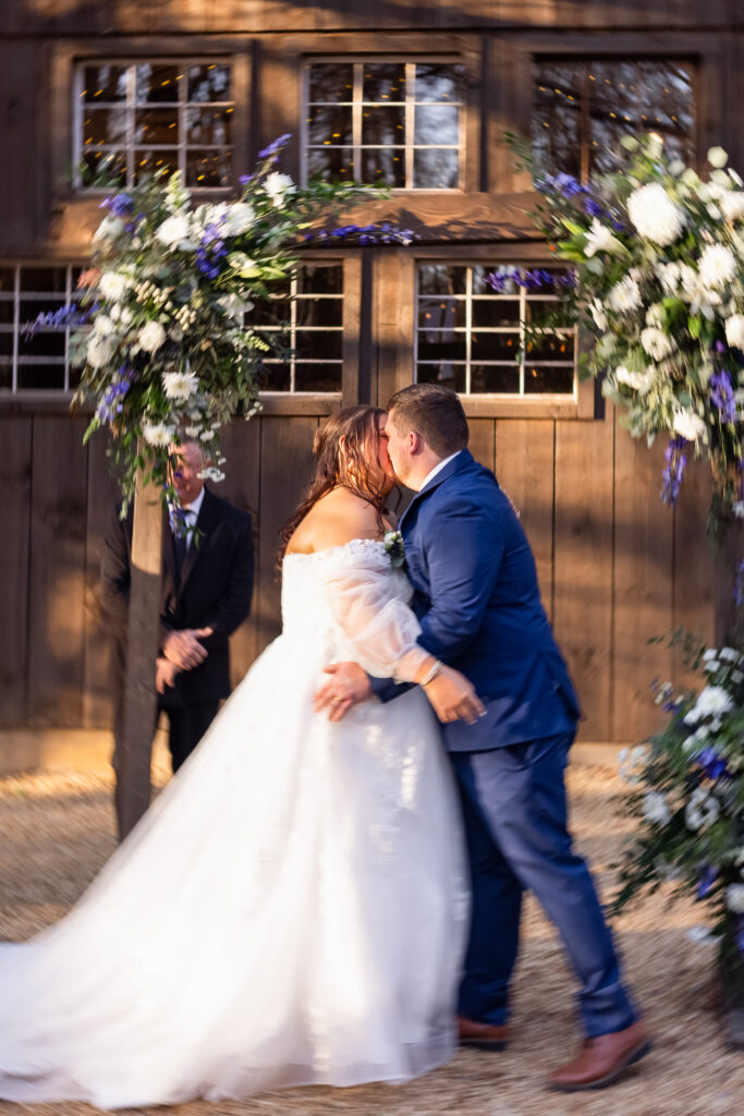 The bride and groom share their first kiss as newlyweds beneath a floral ceremony arch. The groom wears a blue suit while the bride’s wedding dress flows as they lean into the kiss, marking the official conclusion of the wedding ceremony.