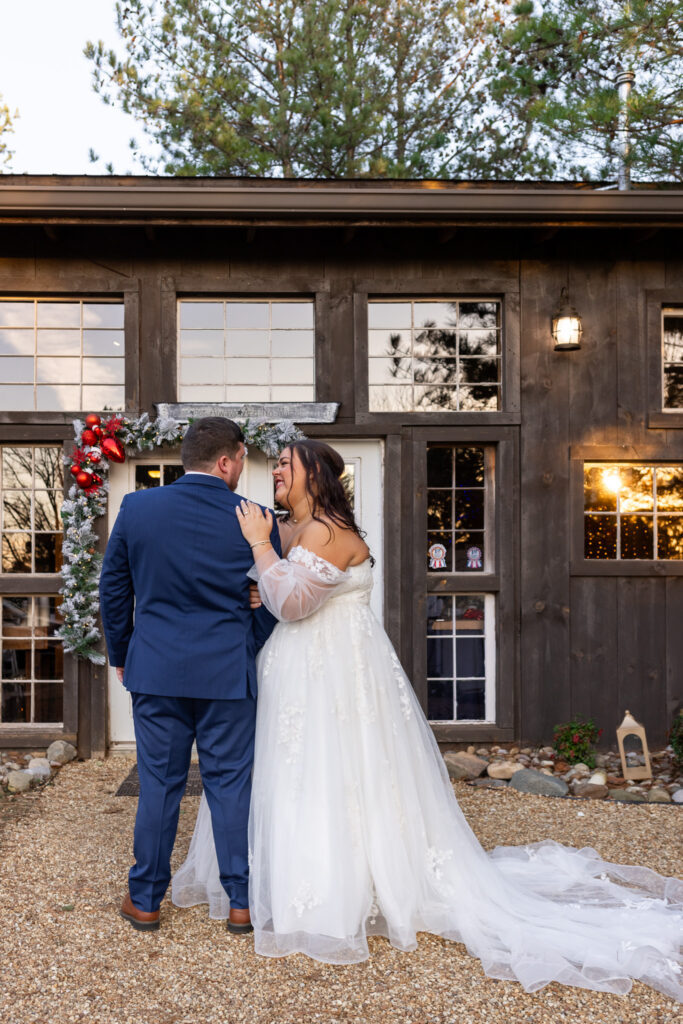 The bride and groom stand close together outside the barn, smiling at one another as the bride rests her hand on the groom’s shoulder during their chattanooga barn wedding, with dark wooden siding, white-trimmed windows, and festive greenery framing the entrance behind them.