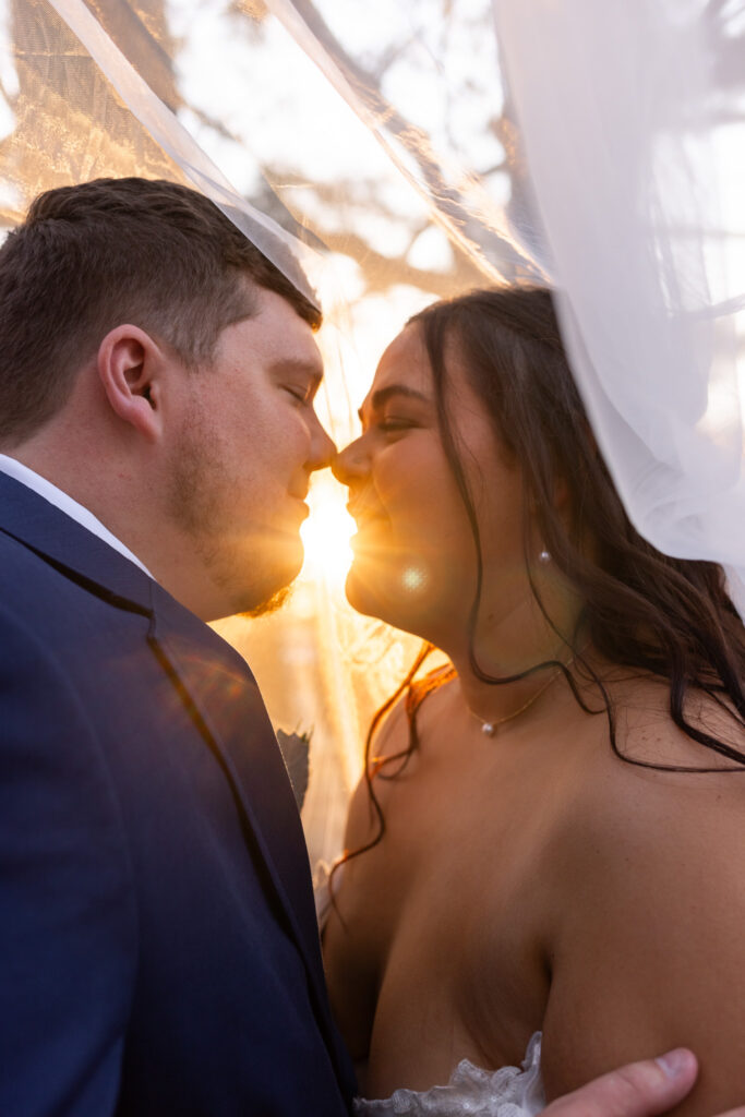 A romantic sunset wedding portrait shows the bride and groom leaning in close beneath the bride’s veil at coffey barn, with golden sunlight flaring between them as they share a quiet newlywed moment.
