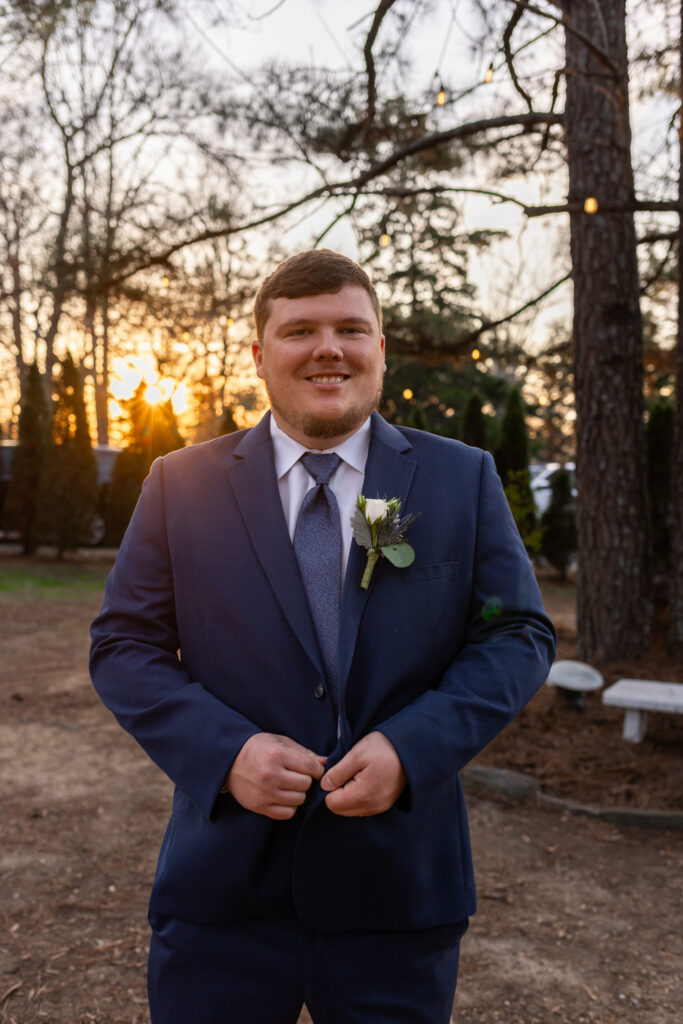 The groom poses alone for a wedding portrait at coffey barn, wearing a blue suit and boutonniere as the sun sets behind the trees, string lights glowing softly in the background.