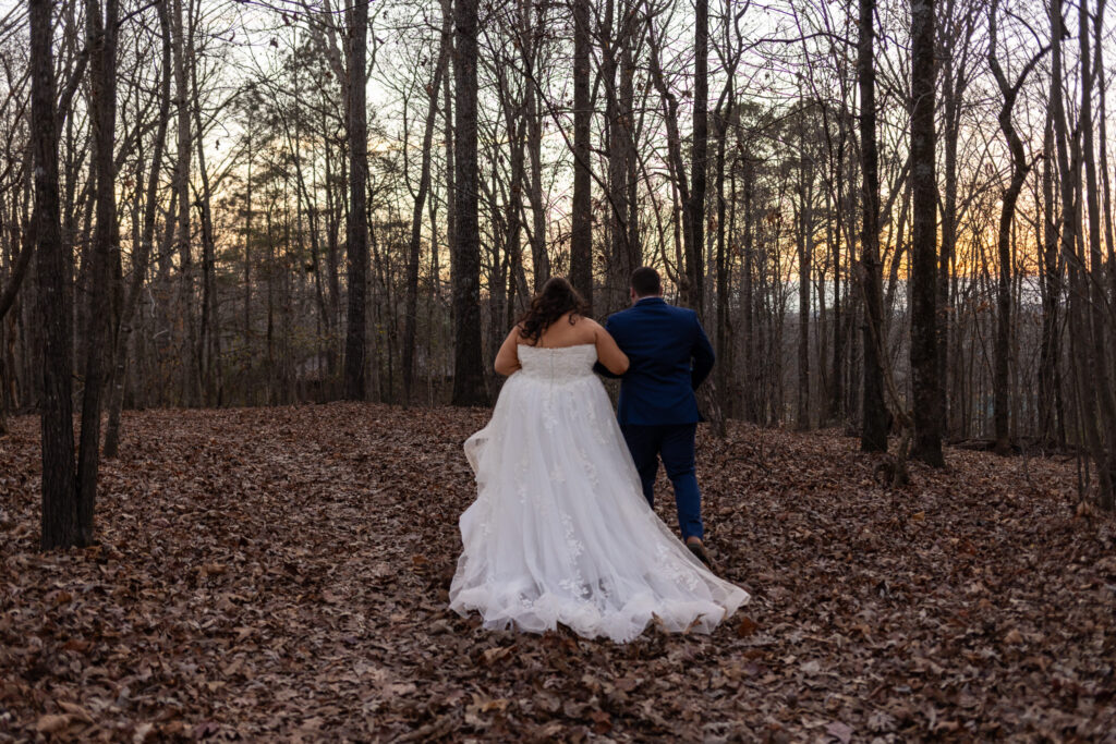 The bride and groom walk away together down a leaf-covered path during their chattanooga barn wedding, viewed from behind as the bride’s dress trails across the forest floor and the sky glows with fading sunset light.